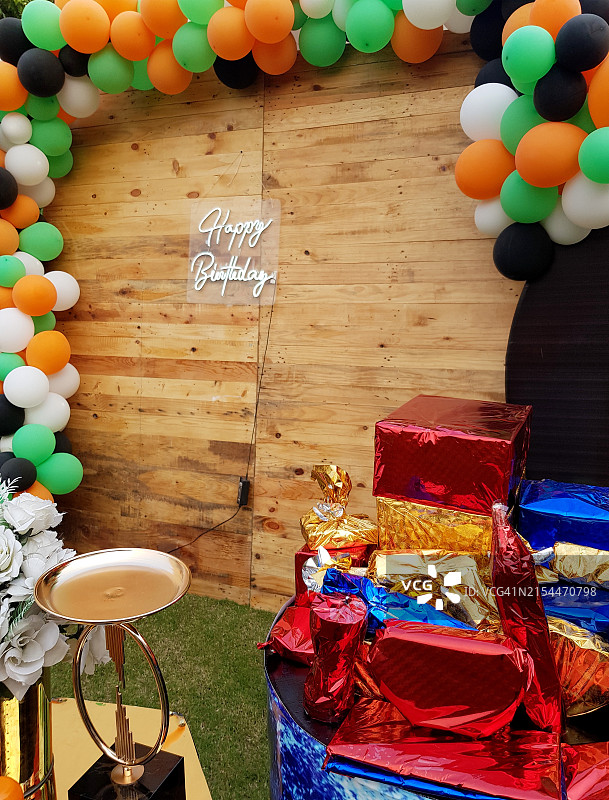 生日快乐，写在木制墙上，墙上装饰着多色气球，前景是用闪亮纸张包裹的生日礼物图片素材