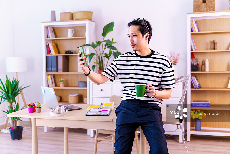 穿条纹衬衫的自信亚洲年轻男人在家办公室内看手机。远程工作，自由职业者，小企业概念图片素材