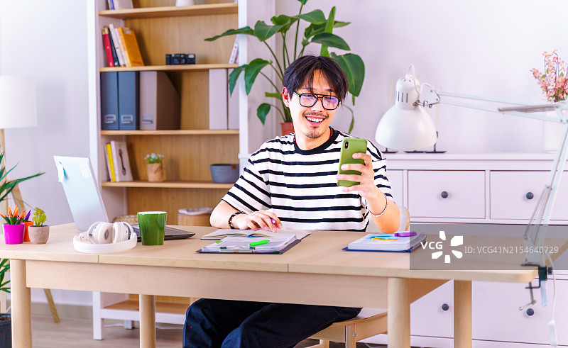年轻自信的亚洲男子身穿条纹衬衫，在家用笔记本电脑工作时看手机。远程办公，自由职业者，小企业概念图片素材