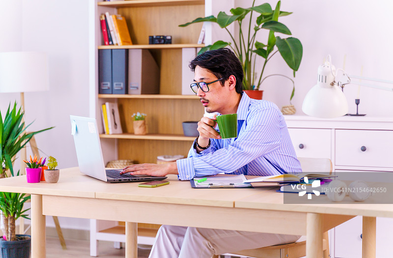 一位穿着蓝色条纹衬衫的自信亚洲年轻男子在家用笔记本电脑工作。远程办公，自由职业者，小型企业概念图片素材
