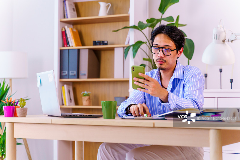 一位自信的亚洲年轻男人穿着蓝色条纹衬衫，在家居办公室里一边用笔记本电脑工作一边看手机。远程办公，自由职业者，小企业概念图片素材