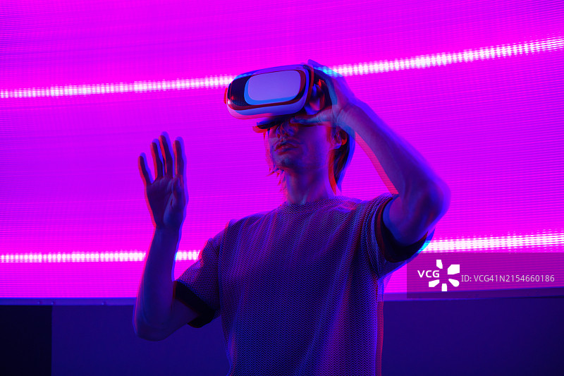 戴着虚拟现实眼镜在蓝色霓虹灯墙背景上手势的惊讶的男人图片素材