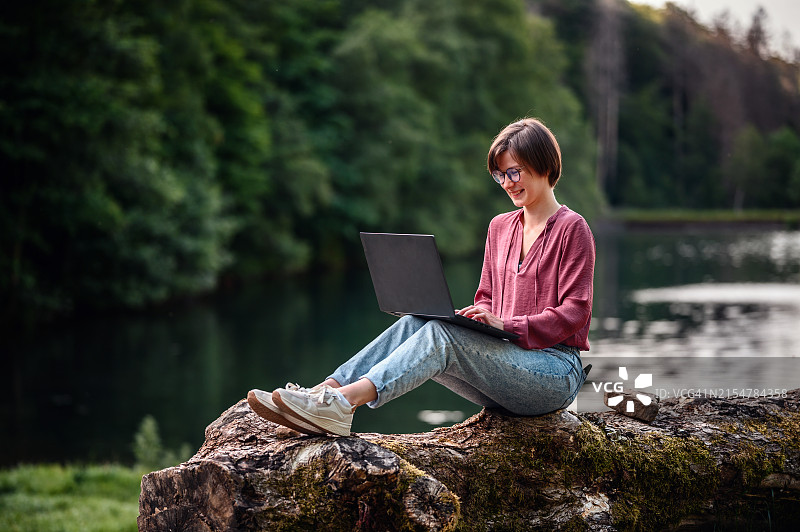 年轻女人在森林里坐在圆木上微笑使用笔记本电脑图片素材