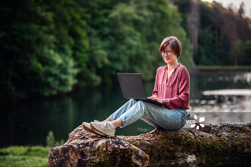 年轻女人在森林里坐在圆木上微笑使用笔记本电脑图片素材