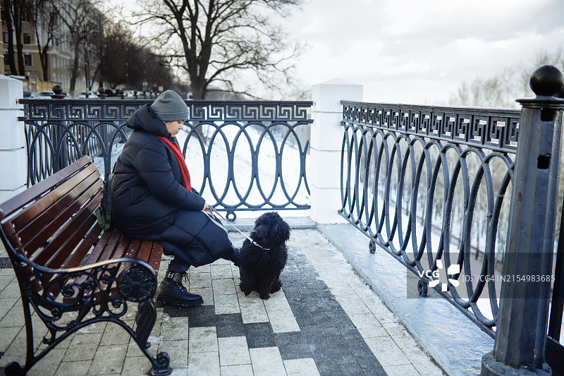 情绪支持：冬季服装的超重女性在城市滨水区与她的狗坐在一起图片素材
