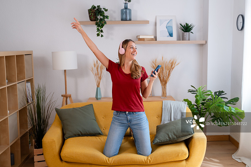 快乐的年轻女子戴着耳机和智能手机在客厅里跳舞图片素材