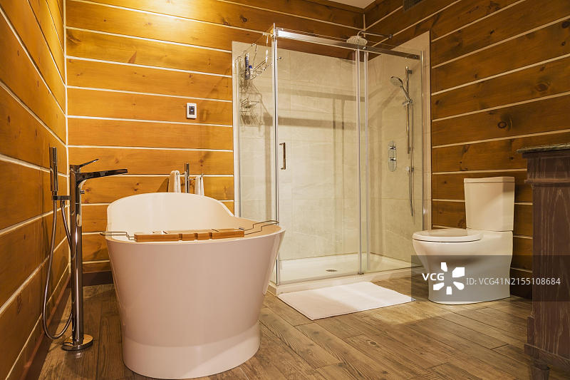 独立式浴缸，带高位水箱马桶和透明玻璃淋浴间，位于套间内，内部铺有仿木瓷砖，位于加拿大魁北克省的白松木和扁平原木房屋内图片素材