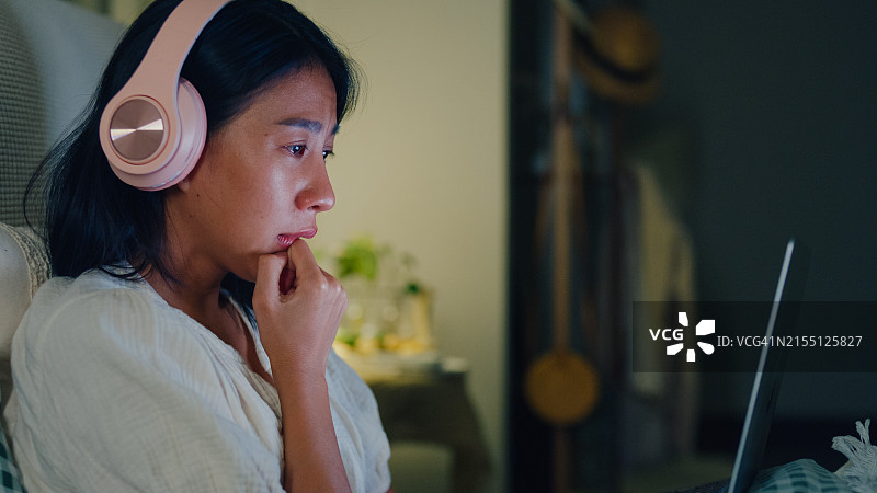 年轻亚洲女孩在床上戴着耳机哭泣，观看流媒体在线视频点播电影特写。失眠、网络病、睡眠障碍图片素材