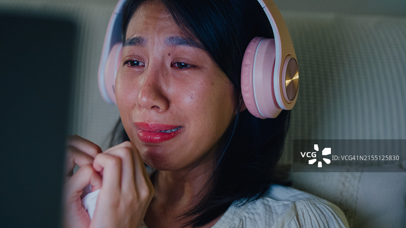 年轻亚洲女孩在床上戴着耳机哭泣，观看流媒体在线视频点播电影特写。失眠、网络病、睡眠障碍图片素材