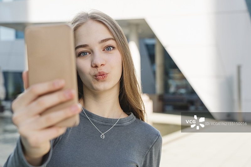 在建筑物前用智能手机嘟嘴自拍的少女图片素材