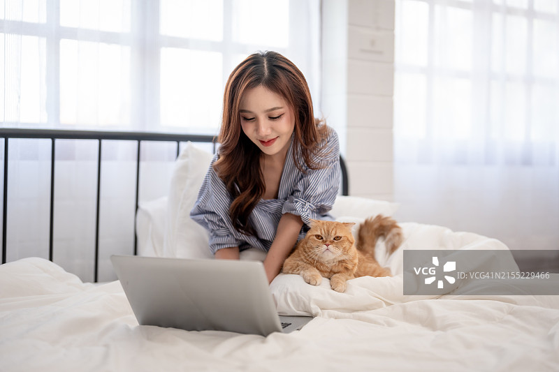 美丽的亚洲女性在家里用笔记本电脑和她的家猫一起工作。有魅力的年轻女宠物主人坐在床上，在卧室里用设备和她的宠物打发休闲时光图片素材
