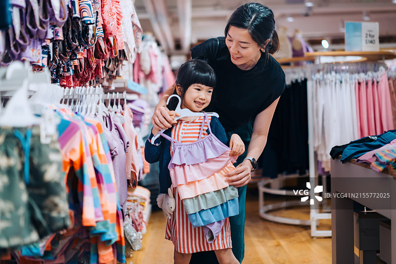 年轻的亚洲母亲在服装店的镜子前为她可爱的小女儿试穿衣服，一起享受购物的乐趣。家庭购物。在购物中心共度家庭时光。零售和消费图片素材