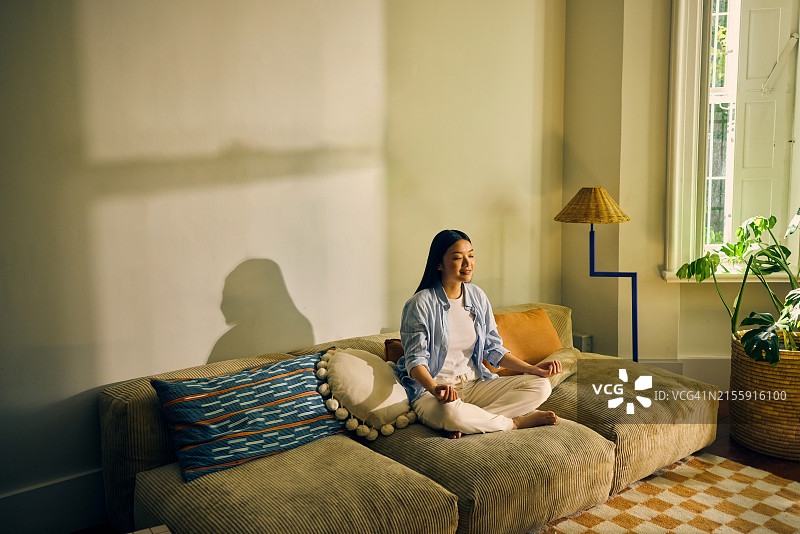 年轻亚裔女性在客厅沙发上冥想，室内生活方式，自我关怀和正念概念图片素材