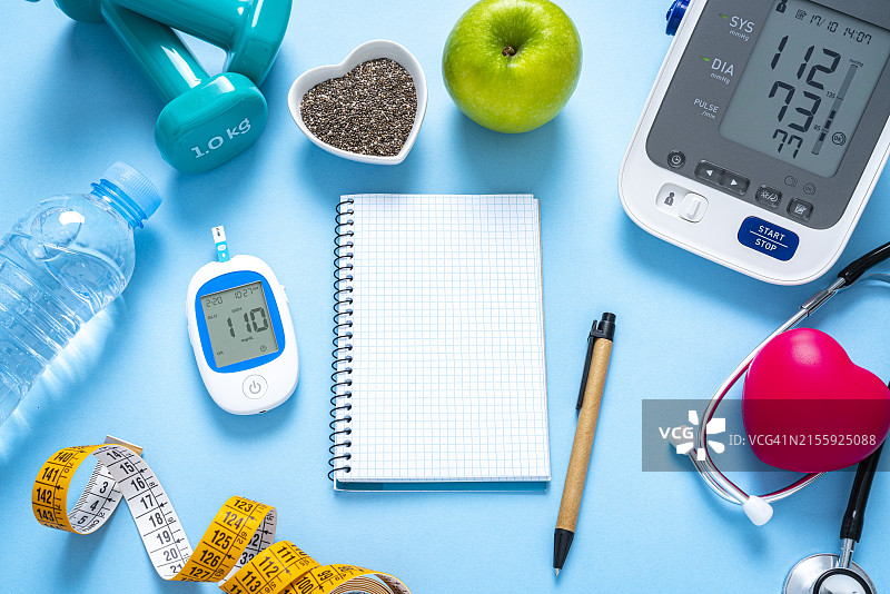 血糖和血压控制概念：血糖仪、血压计、哑铃、青苹果、卷尺，蓝色背景，记事本留白图片素材