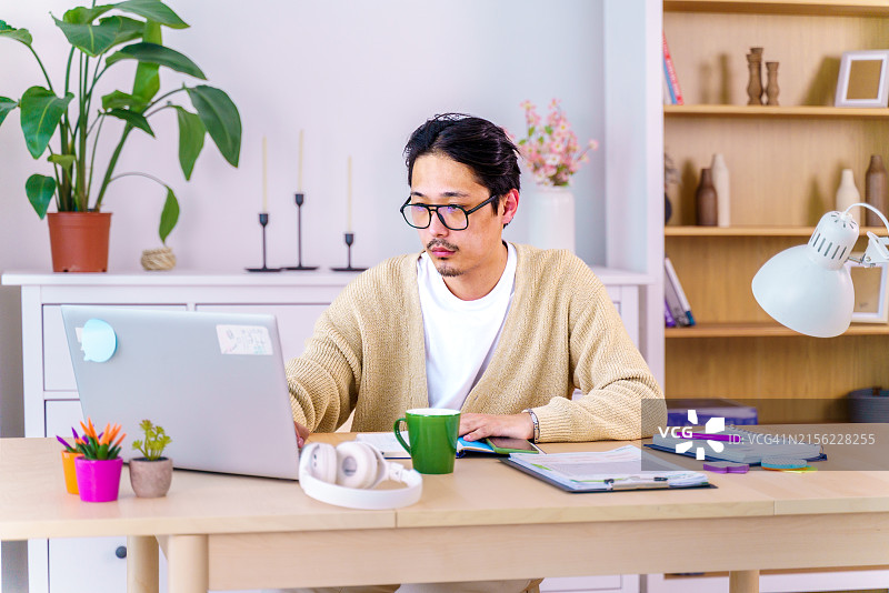 在家庭办公室里用笔记本电脑工作的穿着白色T恤和黄色开衫的自信亚洲青年男子，远程工作，自由职业，小企业概念图片素材