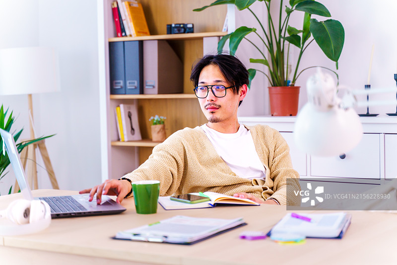 在家庭办公室里用笔记本电脑工作的穿着白色T恤和黄色开衫的自信亚洲青年男子，远程工作，自由职业，小企业概念图片素材