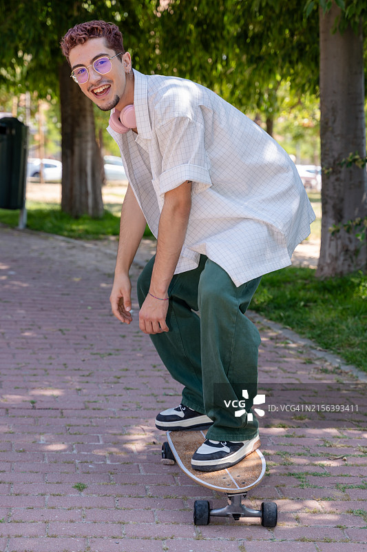 一个面带微笑、留着粉色头发的年轻人，脖子上戴着耳机在公园里玩滑板图片素材