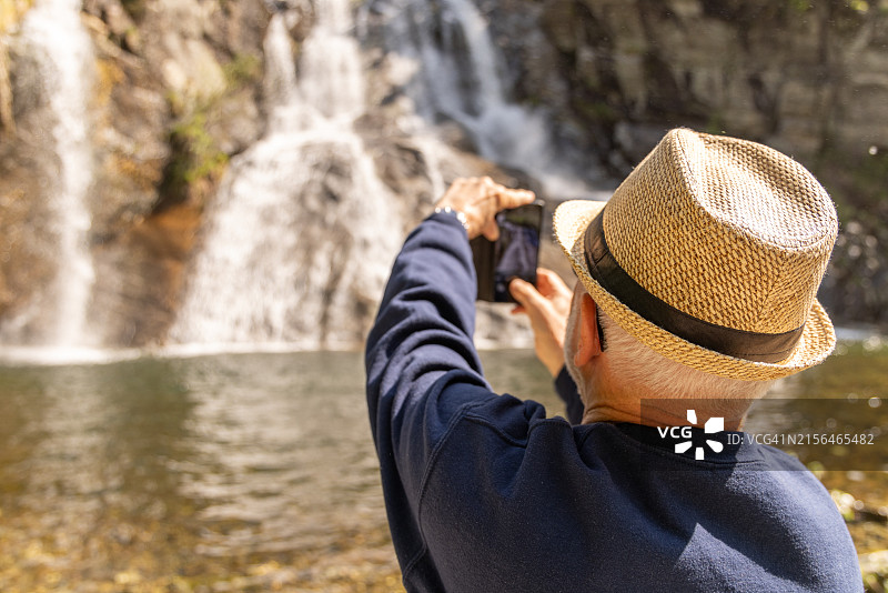 老年男子用智能手机拍摄瀑布背影图片素材