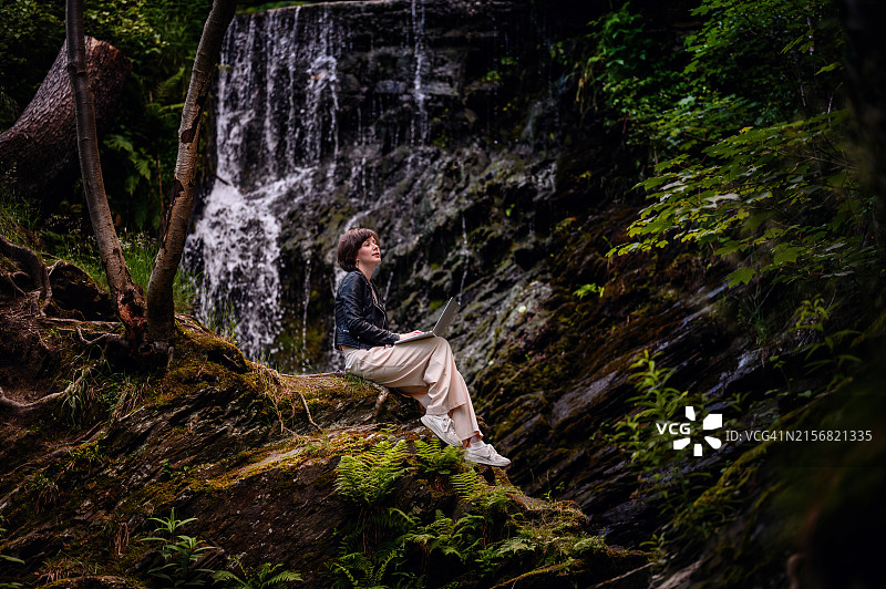 短发皮夹克女人森林瀑布旁用笔记本电脑图片素材