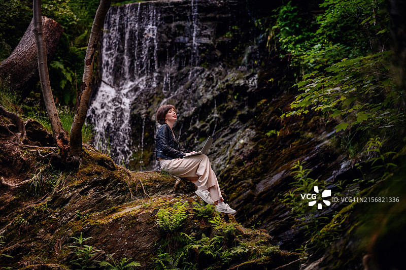 短发皮衣女子在风景如画的瀑布附近的苔藓岩石上用笔记本电脑工作图片素材