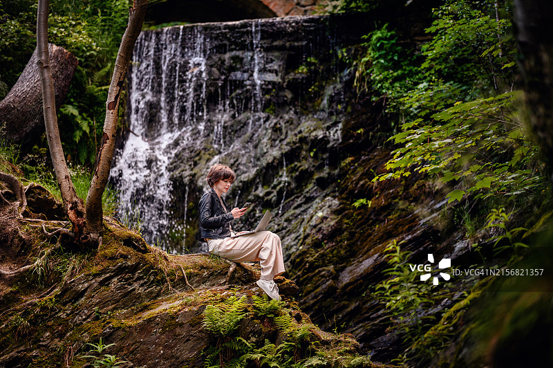 短发皮夹克女人森林瀑布旁用笔记本电脑图片素材