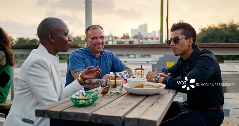 三位朋友在户外露台享受晚餐，夕阳下远处是休斯顿天际线。图片素材
