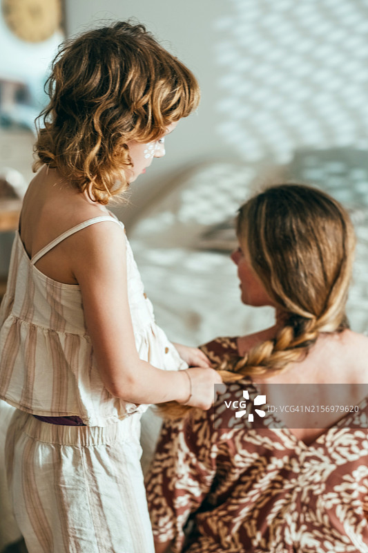 在阳光明媚的卧室里，可爱的女孩为妈妈编辫子，捕捉充满爱和关怀的温馨家庭时刻图片素材