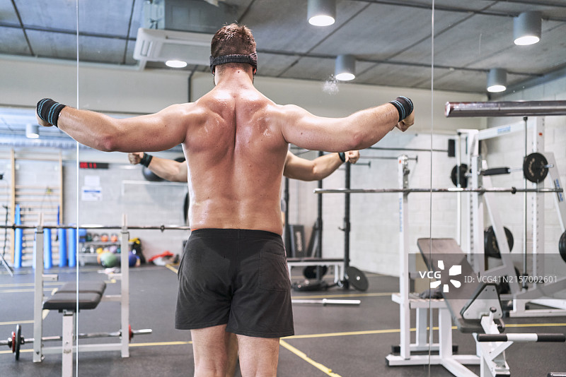 健壮的男人赤裸上身站在健身房里，看着镜子里的自己的背影图片素材