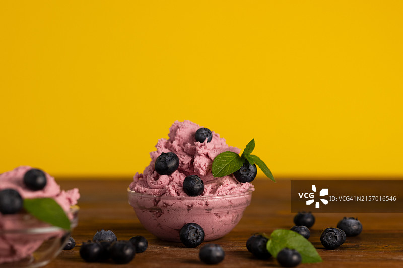 蓝莓装饰玻璃碗装红色水果冰淇淋，木桌黄色背景图片素材