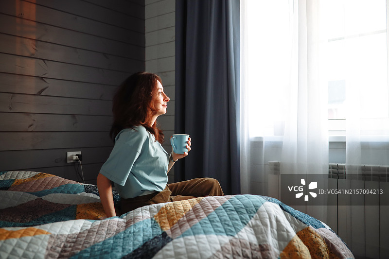 优雅女人坐在卧室的床上，手捧茶杯，透过窗户欣赏日落，展现女性轮廓图片素材