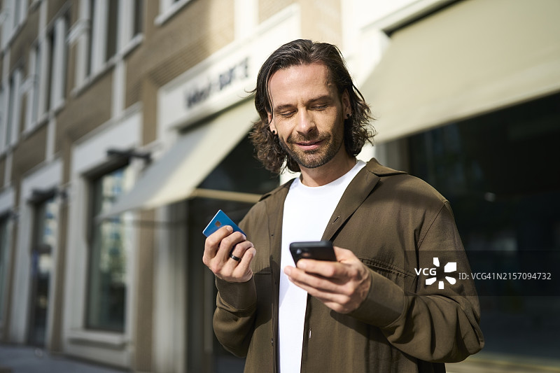 在阳光明媚的日子里，微笑的成熟男人使用智能手机通过信用卡进行网上购物图片素材