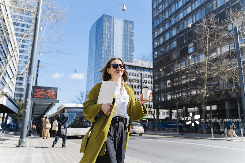 笑容满面的时尚女商人拿着笔记本电脑和一次性杯子走在城市的人行道上图片素材