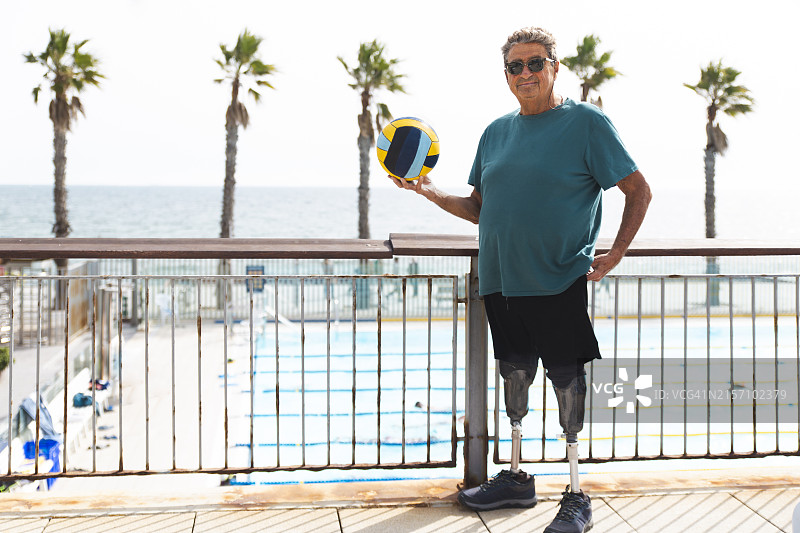 在阳光明媚的日子里，拄着假肢的残疾老人在游泳池附近抱着球图片素材