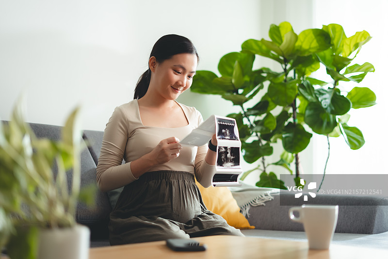亚洲孕妇在家里坐在沙发上，高兴地看着超声波扫描图片素材