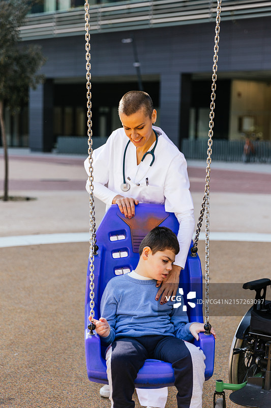 阳光明媚的日子里，护士在公园里推着坐秋千的残疾儿童图片素材