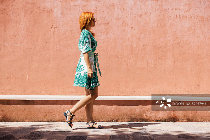 身穿夏装的美丽女人在阳光明媚的日子里走在墙边的石板路上图片素材