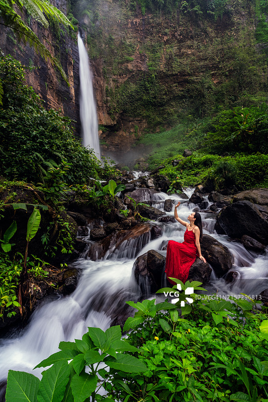 穿着红色连衣裙的女人站在印度尼西亚东爪哇省卡帕斯比鲁瀑布，周围绿树环绕图片素材
