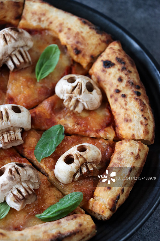 万圣节派对食品特写图像，黑色盘子上切片的玛格丽特披萨，融化的金色水牛奶酪，浓郁的番茄酱，骷髅设计蘑菇和新鲜罗勒叶配料，食物雕刻，眼窝，牙齿，灰色背景图片素材