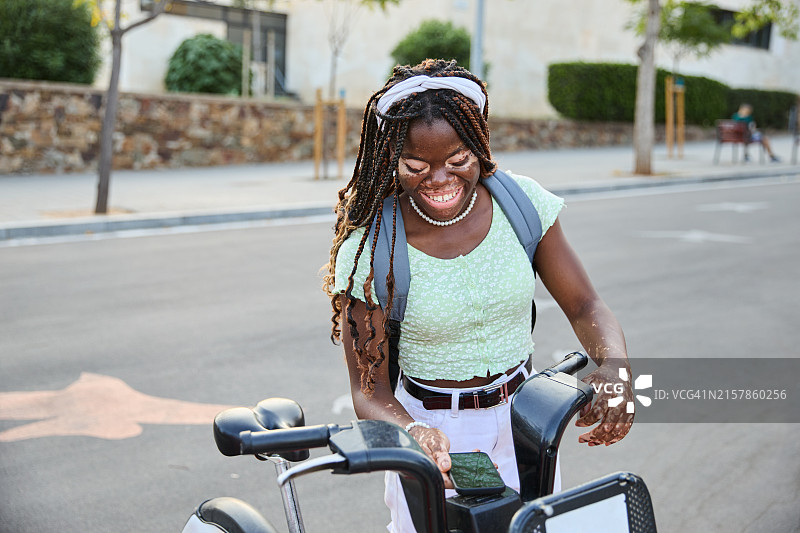 患有白癜风的年轻黑人女学生在城市的自行车租赁站通过智能手机租用电动自行车。环保交通概念图片素材
