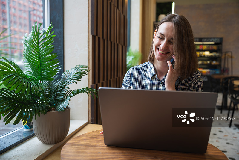 一位女士在咖啡馆靠窗的位置使用笔记本电脑，边打电话边笑着交谈图片素材