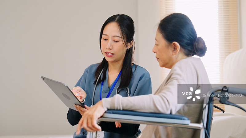 亚洲护理人员护士穿着蓝色工作服向老年退休妇女解释医疗程序和医疗记录，平板电脑上的应用，技术在患者护理中的整合以及医疗保健环境中清晰沟通的重要性图片素材