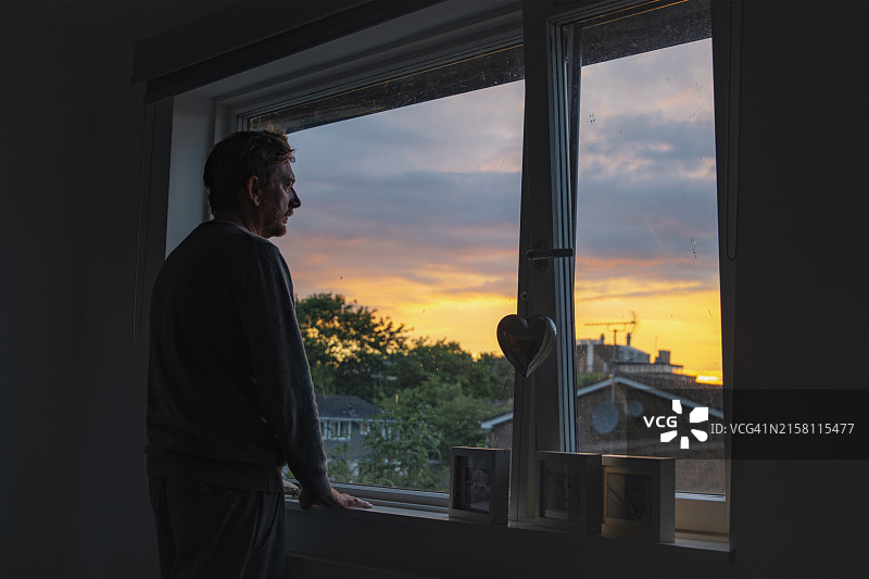 一位沉思的中年男人在卧室的窗边看日落图片素材