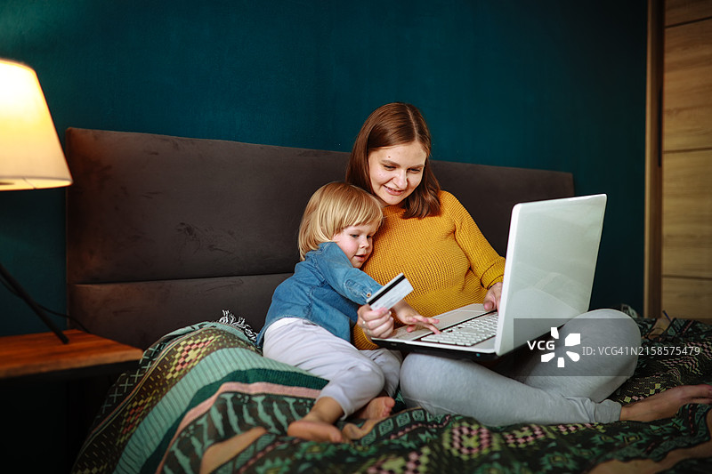 母亲和幼儿儿子在床上使用笔记本电脑和银行卡进行网上购物图片素材