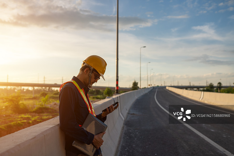戴着黄色安全帽的男人站在桥上，看着他的手机图片素材
