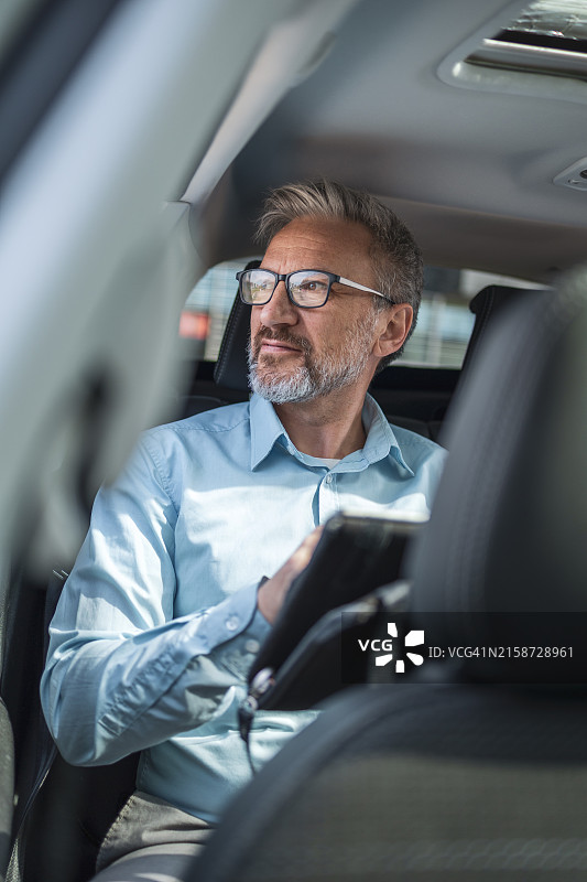 成熟的商务专业人士在出租车上远程工作，戴着眼镜和胡须的男士拿着平板电脑，若有所思地看着窗外，享受着阳光明媚的一天图片素材