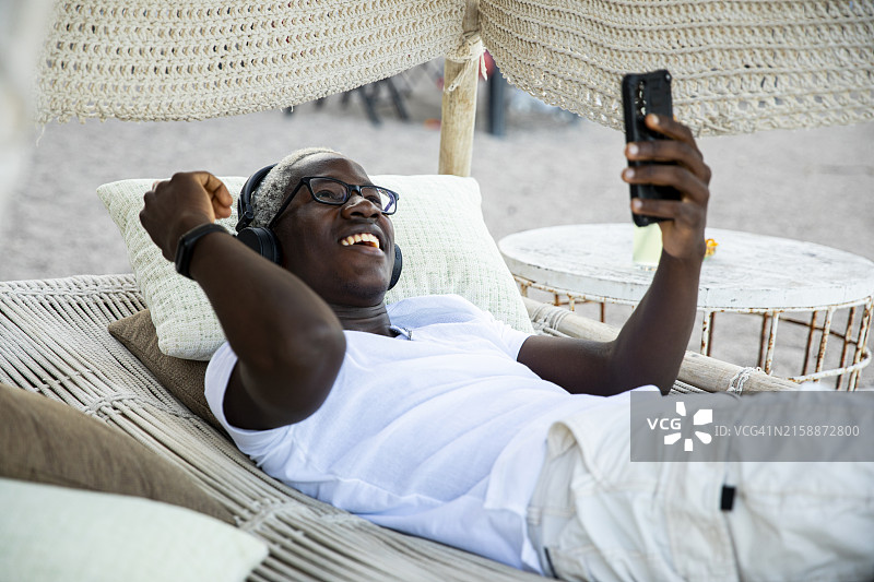 快乐的千禧一代男孩戴着耳机，用手机听音乐，躺在吊床上跳舞图片素材