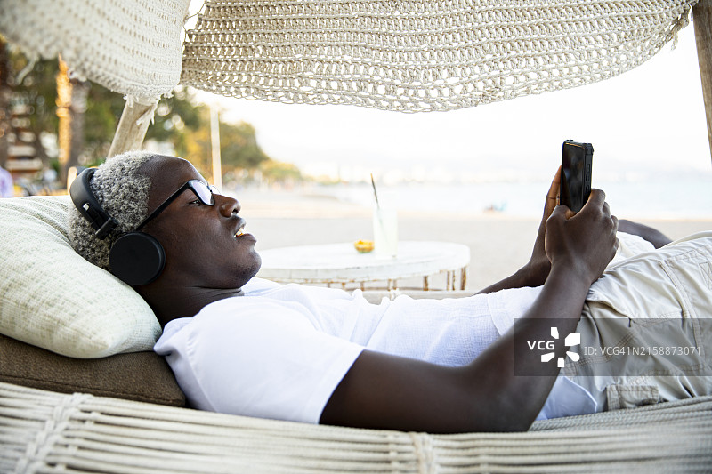 快乐的千禧一代男孩戴着耳机，用手机听音乐，躺在吊床上跳舞图片素材