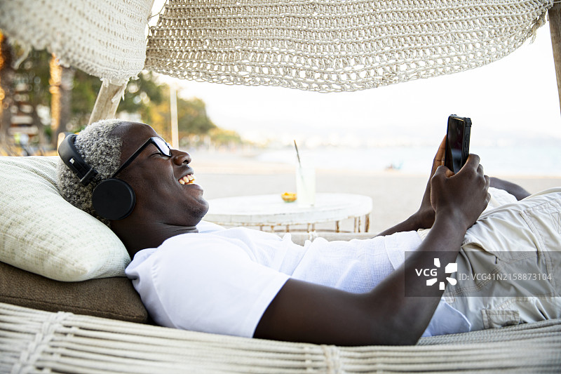 快乐的千禧一代男孩戴着耳机，用手机听音乐，躺在吊床上跳舞图片素材