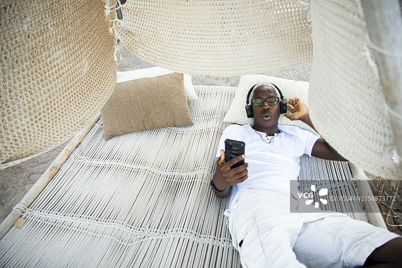 快乐的千禧一代男孩戴着耳机，用手机听音乐，躺在吊床上跳舞图片素材