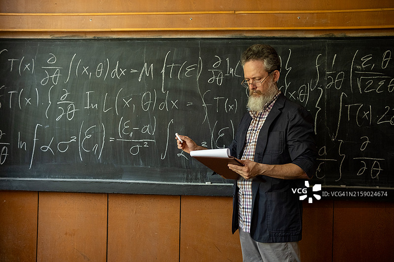 一位留着长白胡子、蓄着大灰发、戴着眼镜的老师，穿着格子衬衫和运动夹克，站在写满复杂公式的黑板前。图片素材